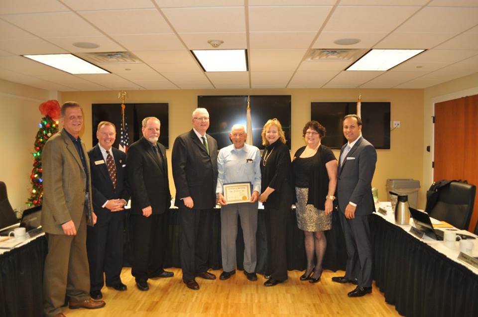 Five men and two women smile and stand with a man in a blue shirt holding a certificate in a room with a Christmas tree in the left corner.