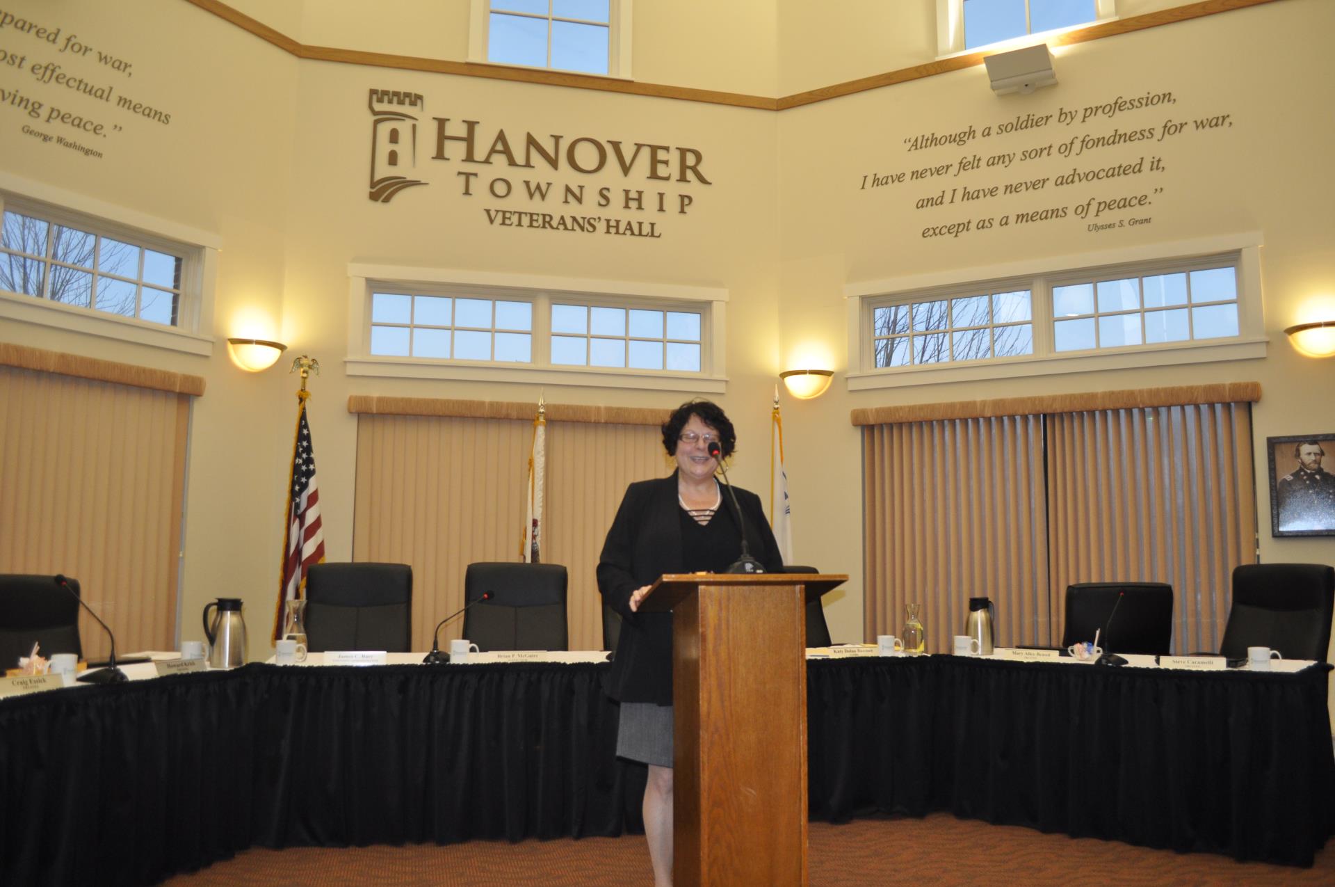 A woman dressed in black with short black hair and glasses stands and presents at a podium speaking into a microphone.