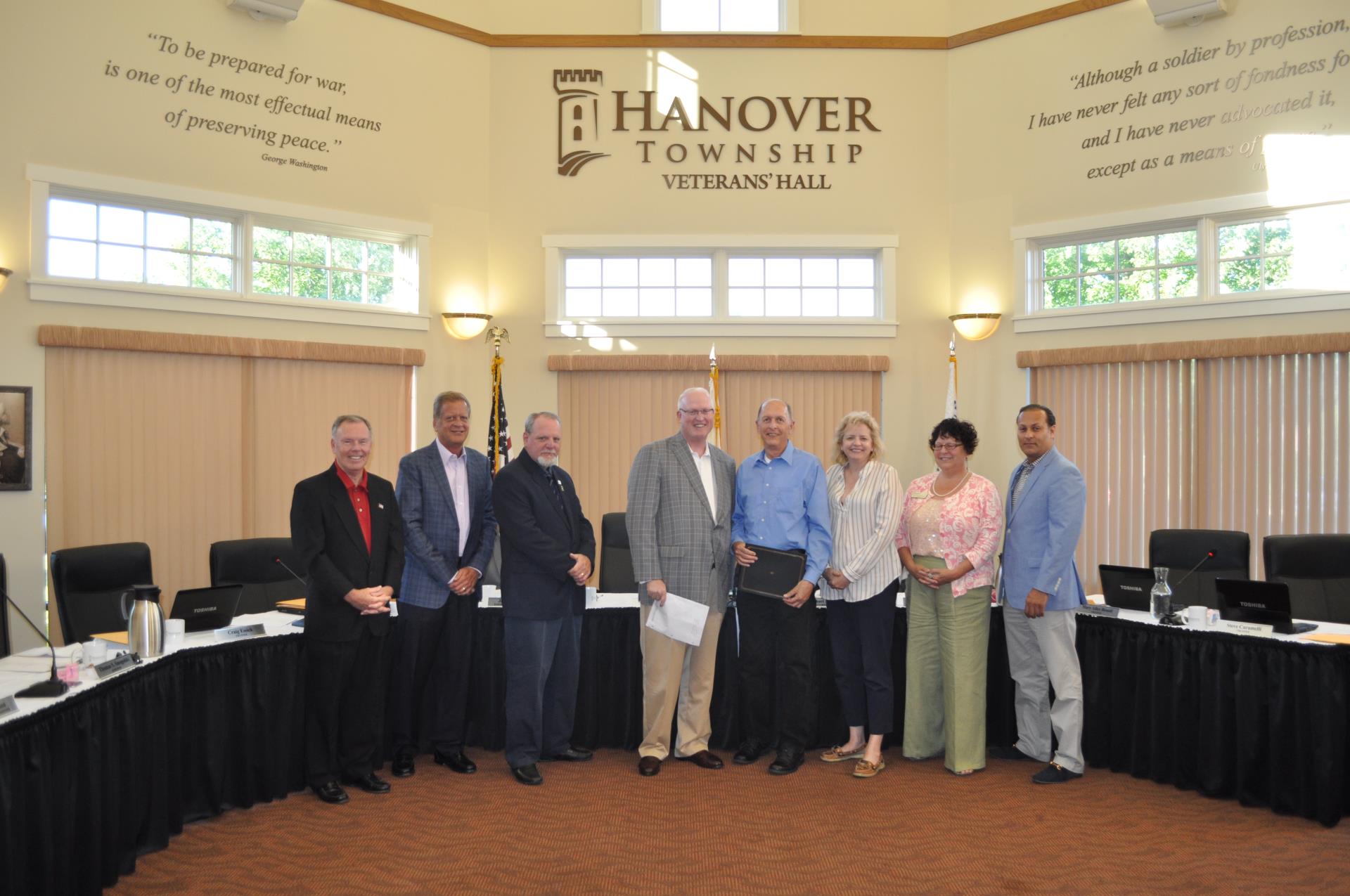 Six men and two women smile for a photo inside of Veterans' Hall. A conference table is behind them.