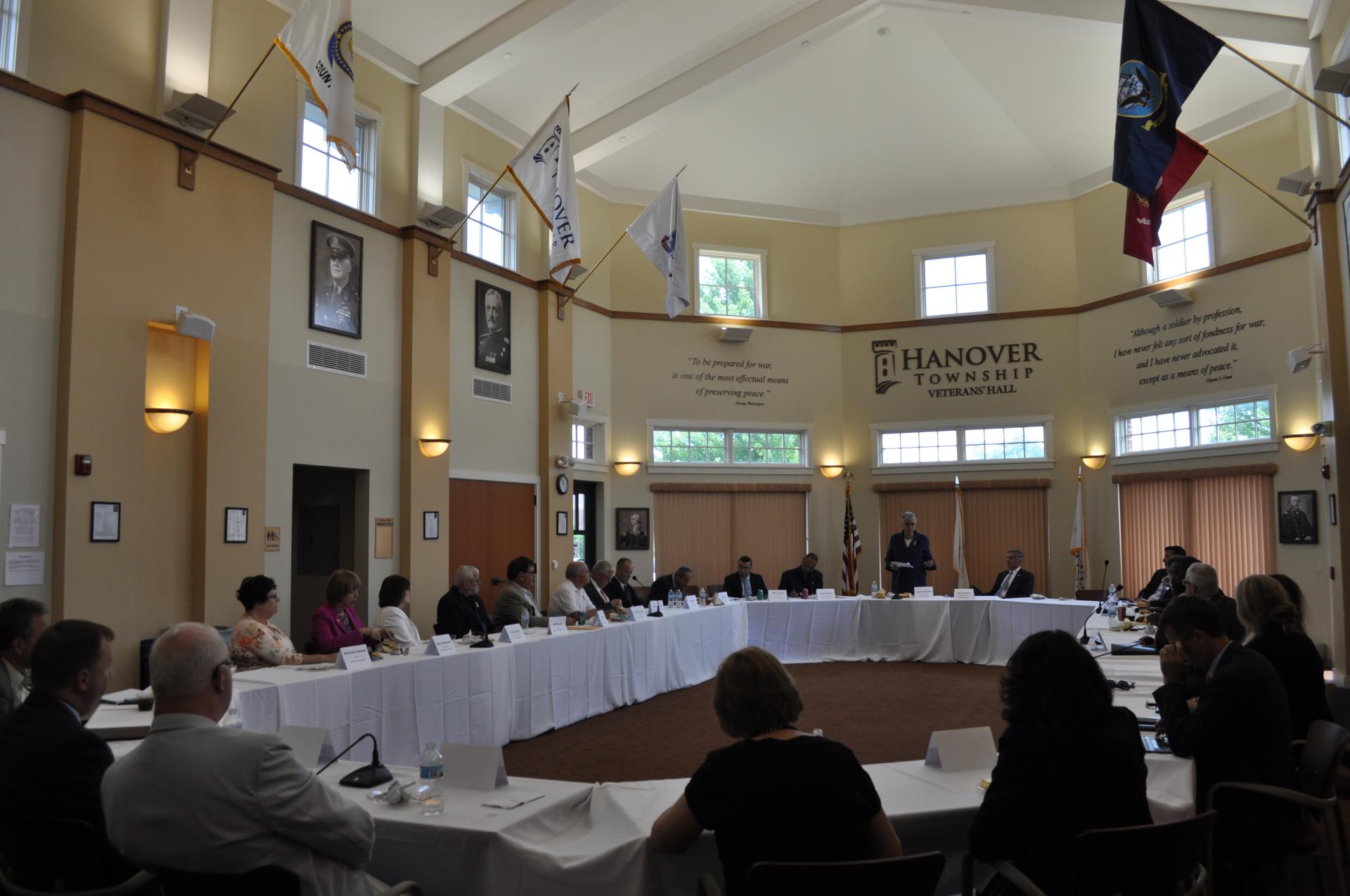 Men and woman sit around a big circle conference table inside bright room with flags on the wall.