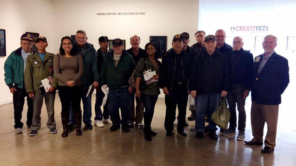 Thirteen men and two women stand and smile together in the middle of an art gallery. 