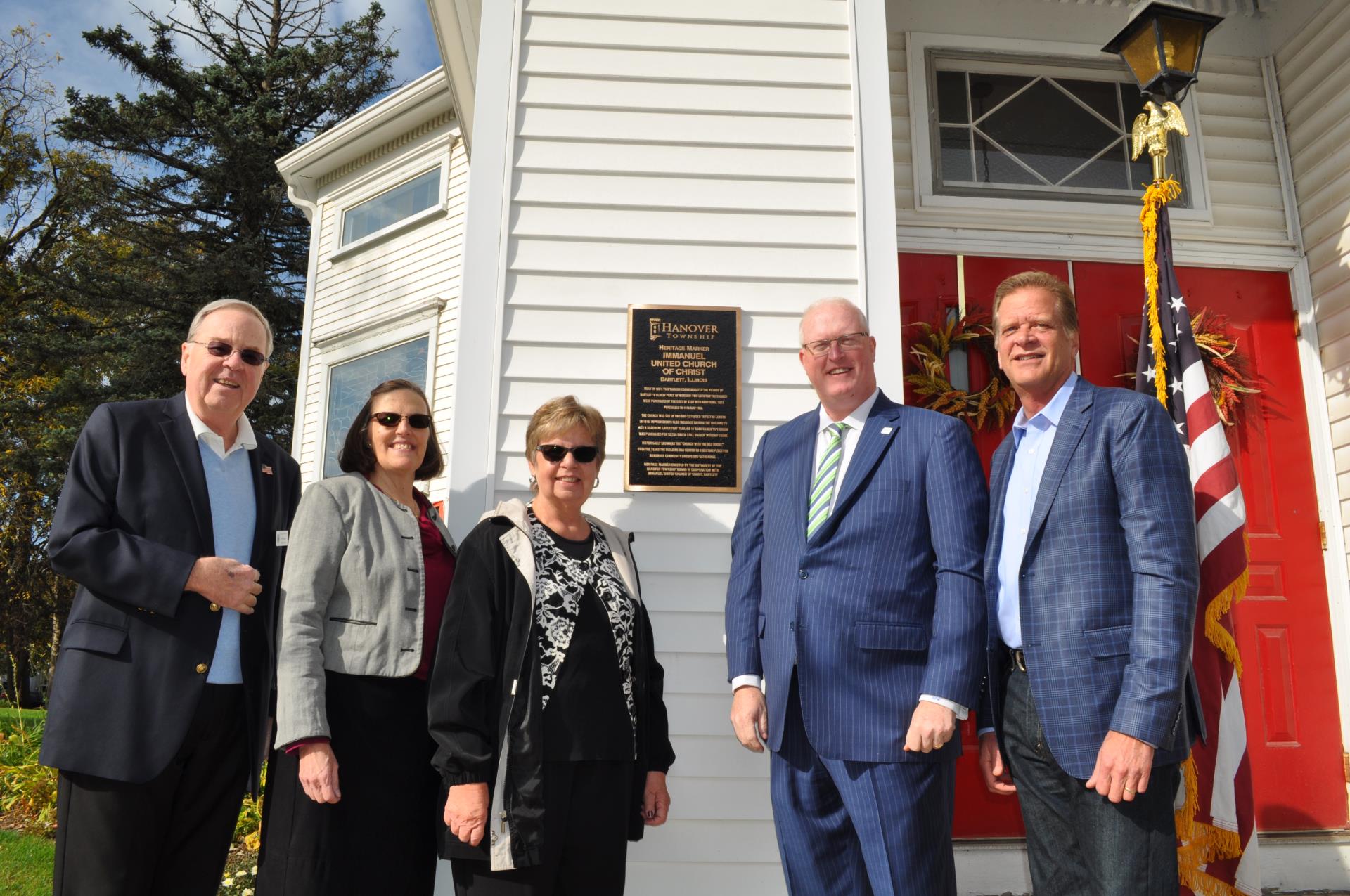 Three men and two women stand next to a black and gold Hanover Township Heritage Marker Immanuel United Church of Christ sign which hangs on the front of the white church. Trees are in the background and it is sunny. 