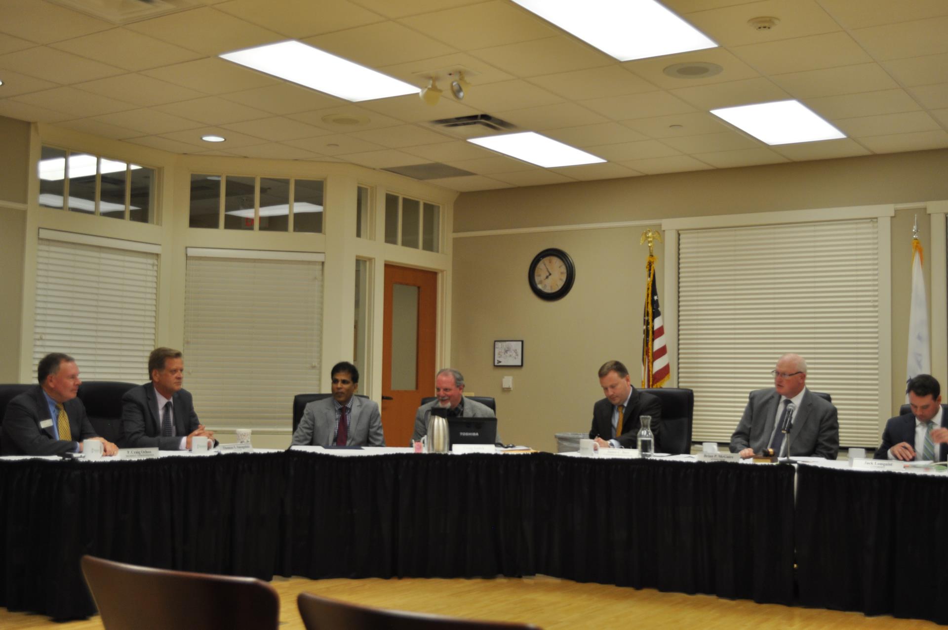 Six men in suits sit around a table at a meeting.