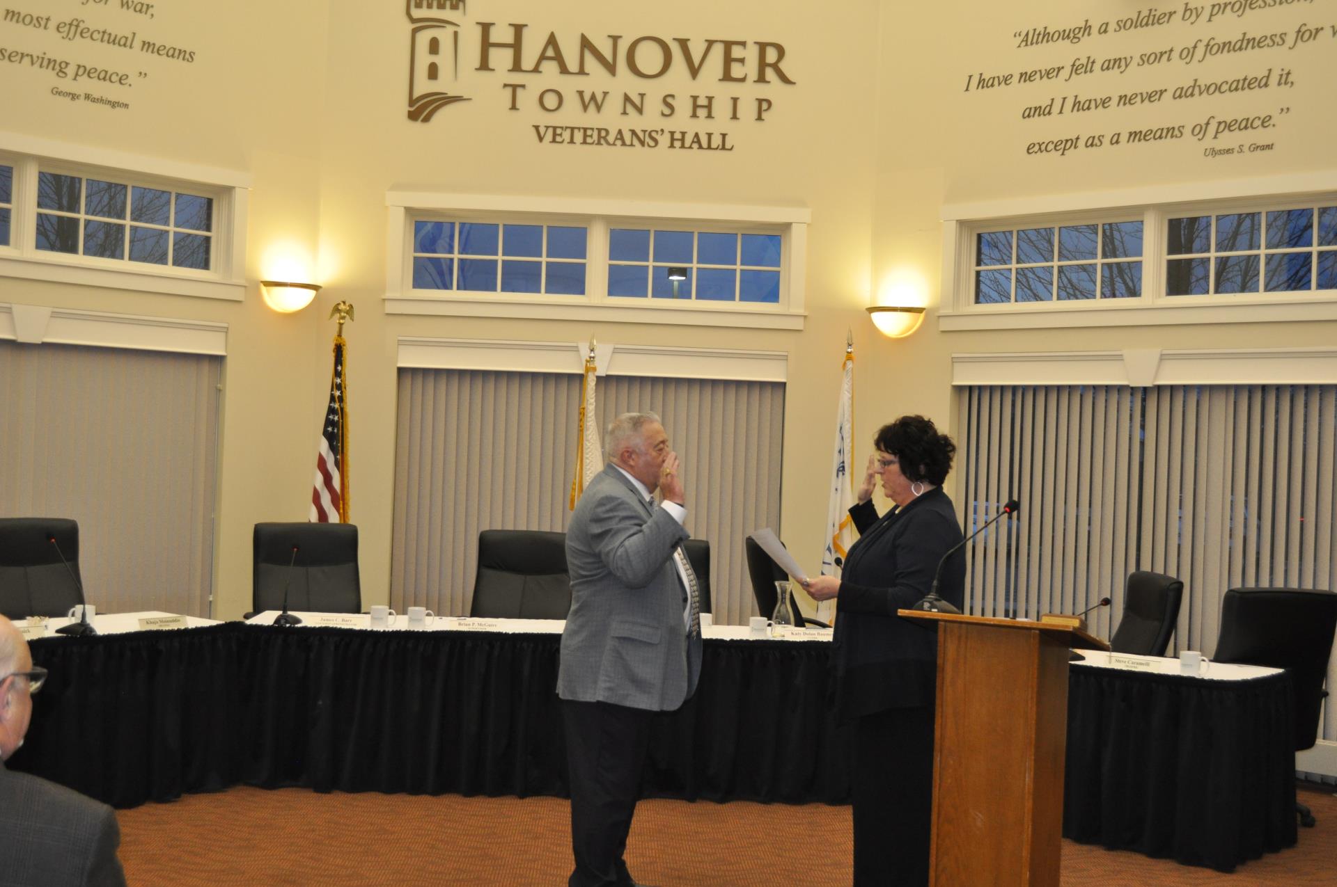 A man in a grey suit holds up his right hand as he gets sworn in by a woman in a dark blue outfit who is holding a paper in her left hand and holding up her right hand.