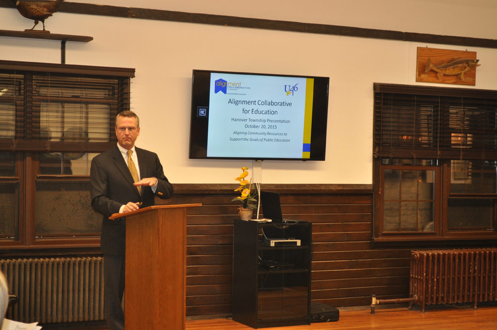 Man in black suit and yellow tie speaks at a podium in a room with two windows and a tv on the wall. 