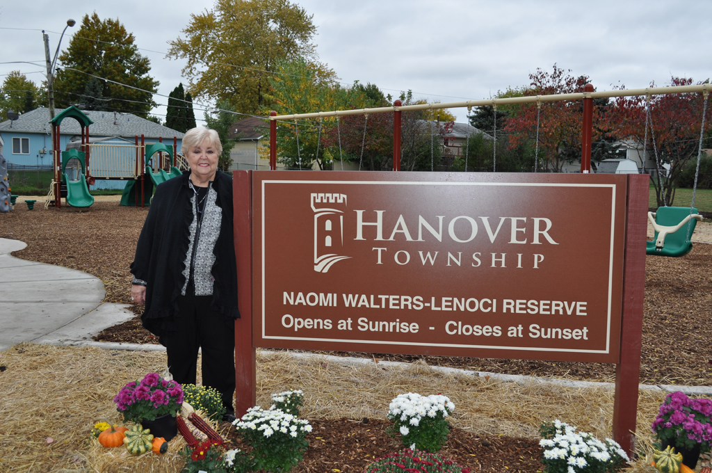 A woman dressed in black and white stands to the left of a brown Hanover Township Naomi Walters- Lenoci Reserve sign that states the park opens at sunrise and closes at sunset. It is cloudy outside.