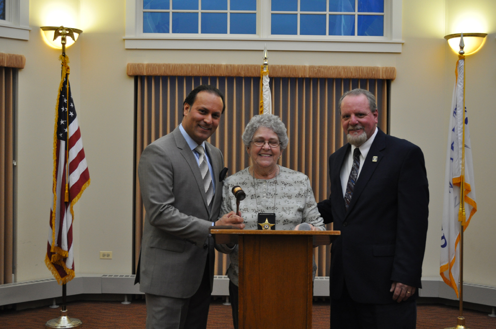 A women with grey hair and glasses wearing a badge around her neck stands and smiles between to men in suits who are also smiling. The three stand in front of a wooden podium with three flags standing behind them.