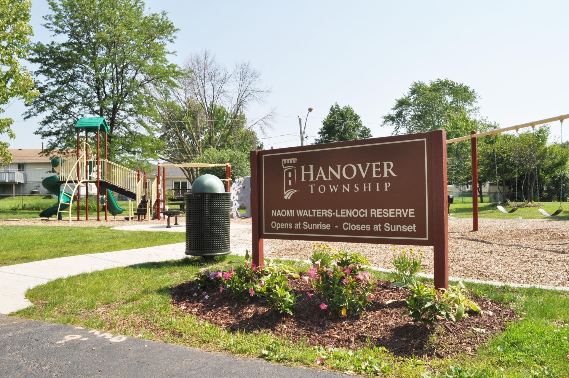 Brown pole graphic sign with the Township logo  and park name in front of the reserve's playground on a sunny day.